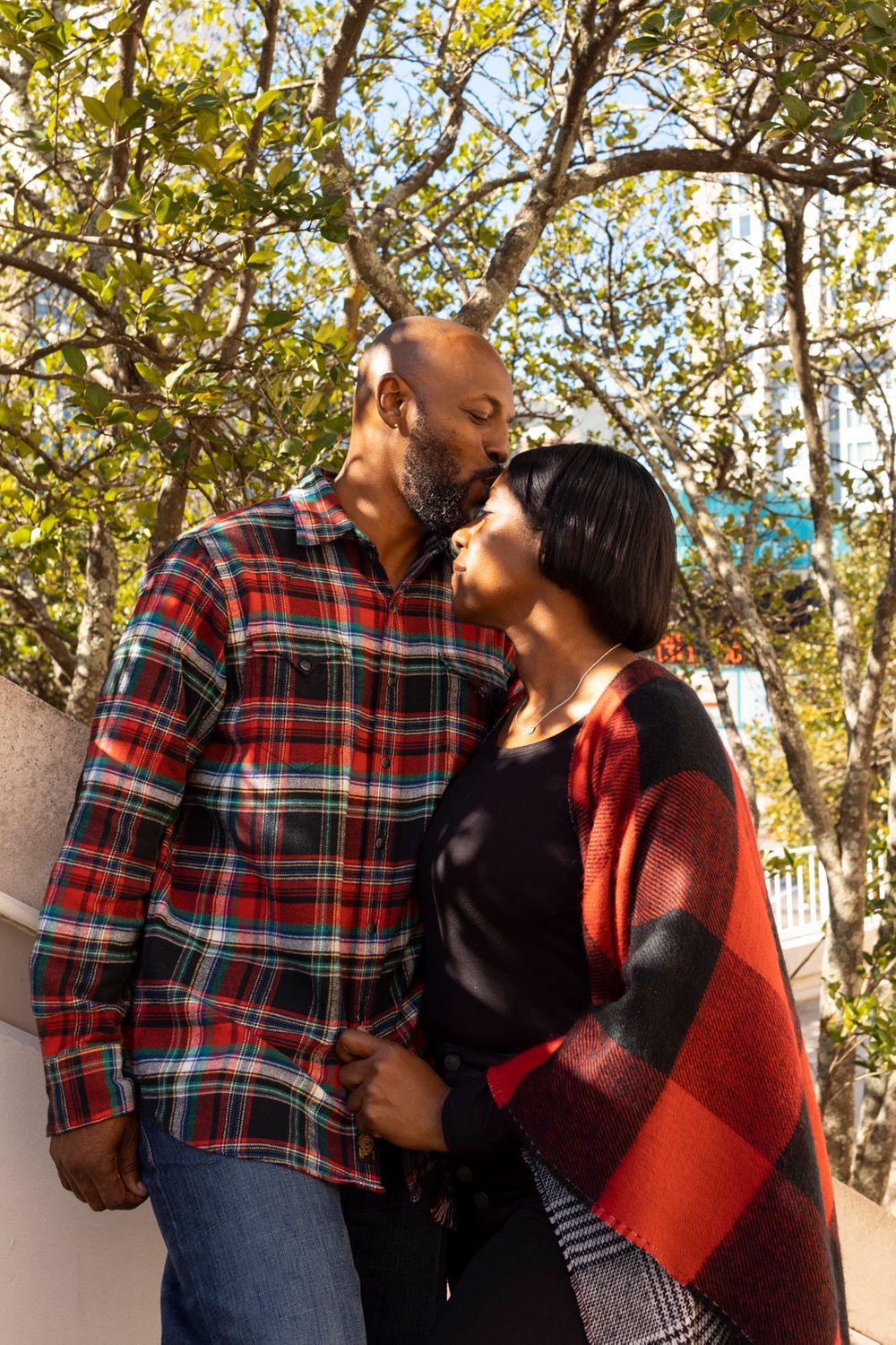 A couple standing under a tree with one person kissing the others forehead.
