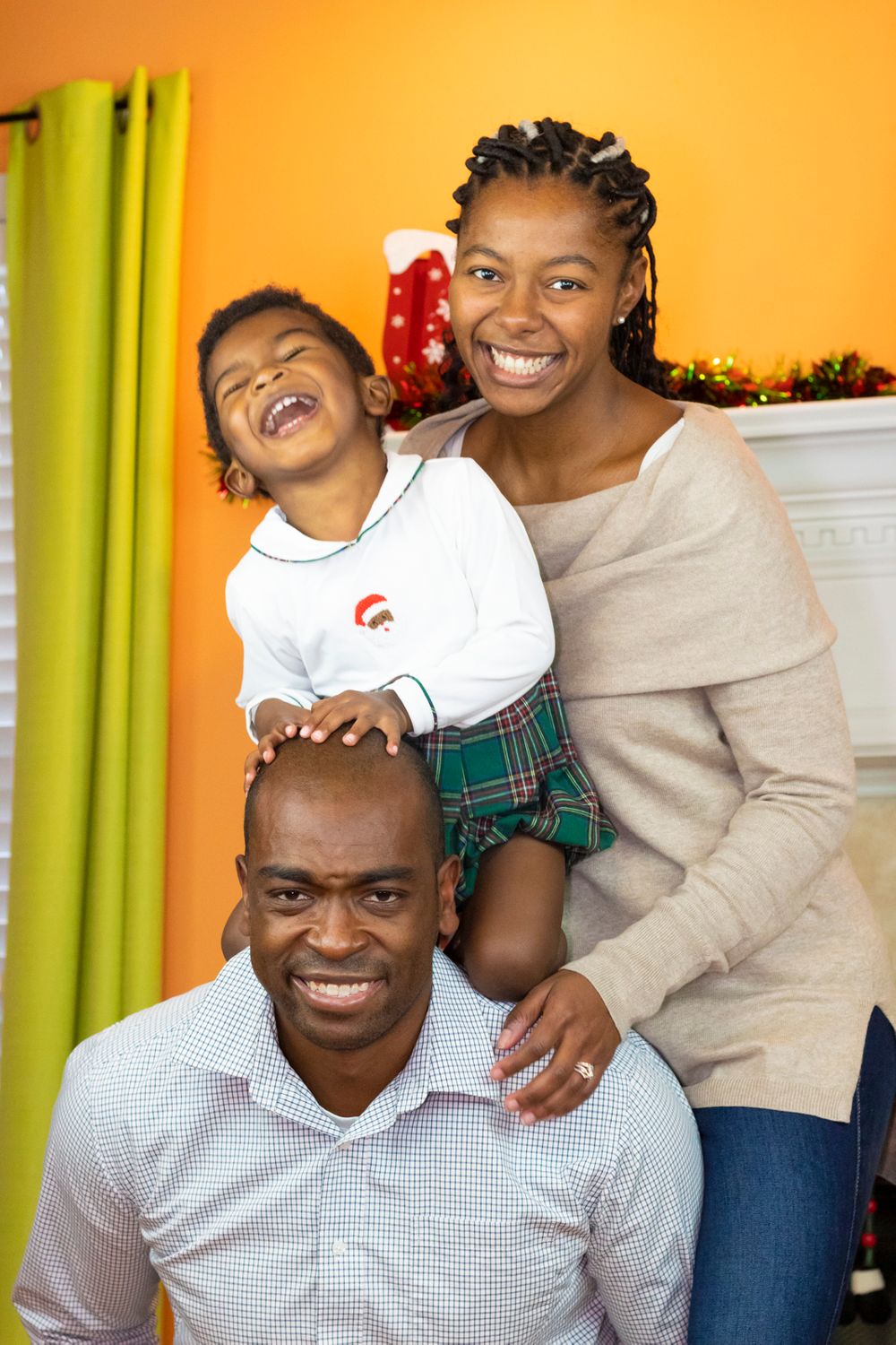 A family of three, the father has the child on his back and the mother is leaning next to them.