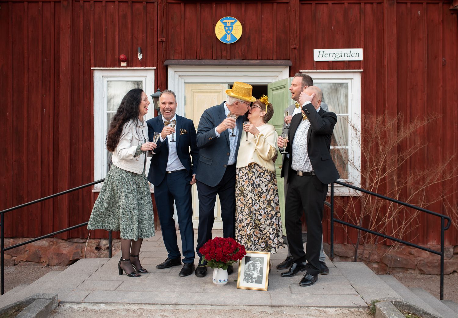 Celebratory gathering of formally dressed people outside red building entrance with festive decor.