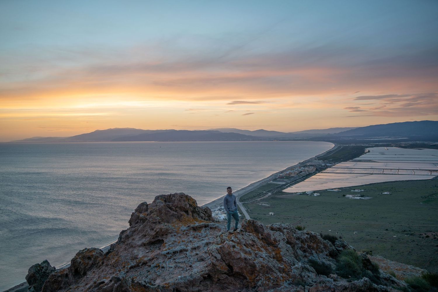 Person standing on cliff overlooking coastal landscape at sunset.