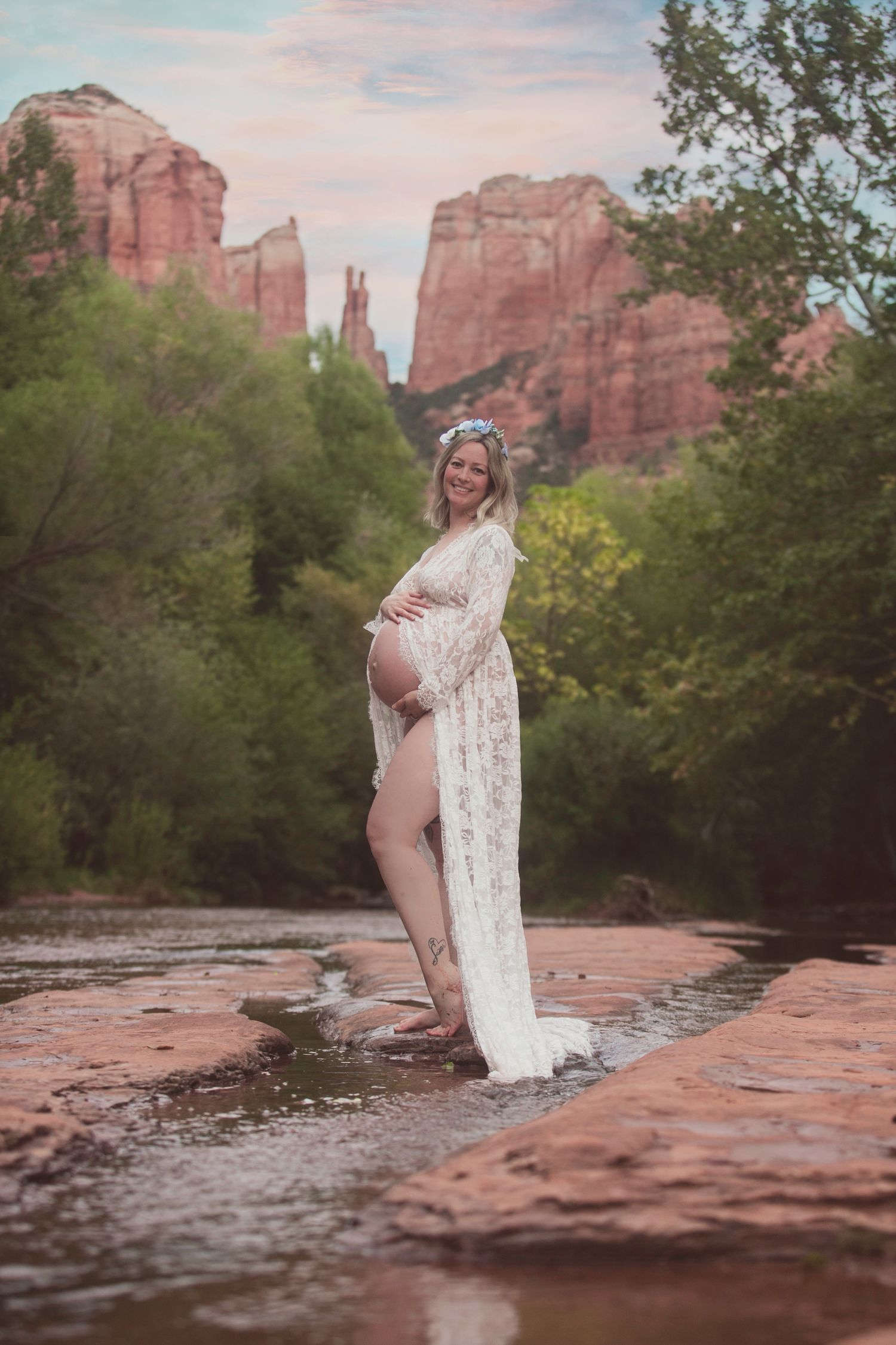 Pregnant woman in white lace maternity gown stands on rocks in Sedona creek with red rock backdrop.