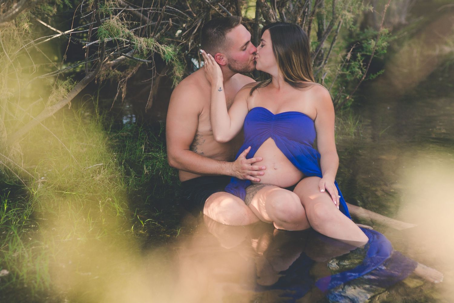 Couple kissing tenderly by a peaceful creek, pregnant woman in elegant blue strapless maternity gown.
