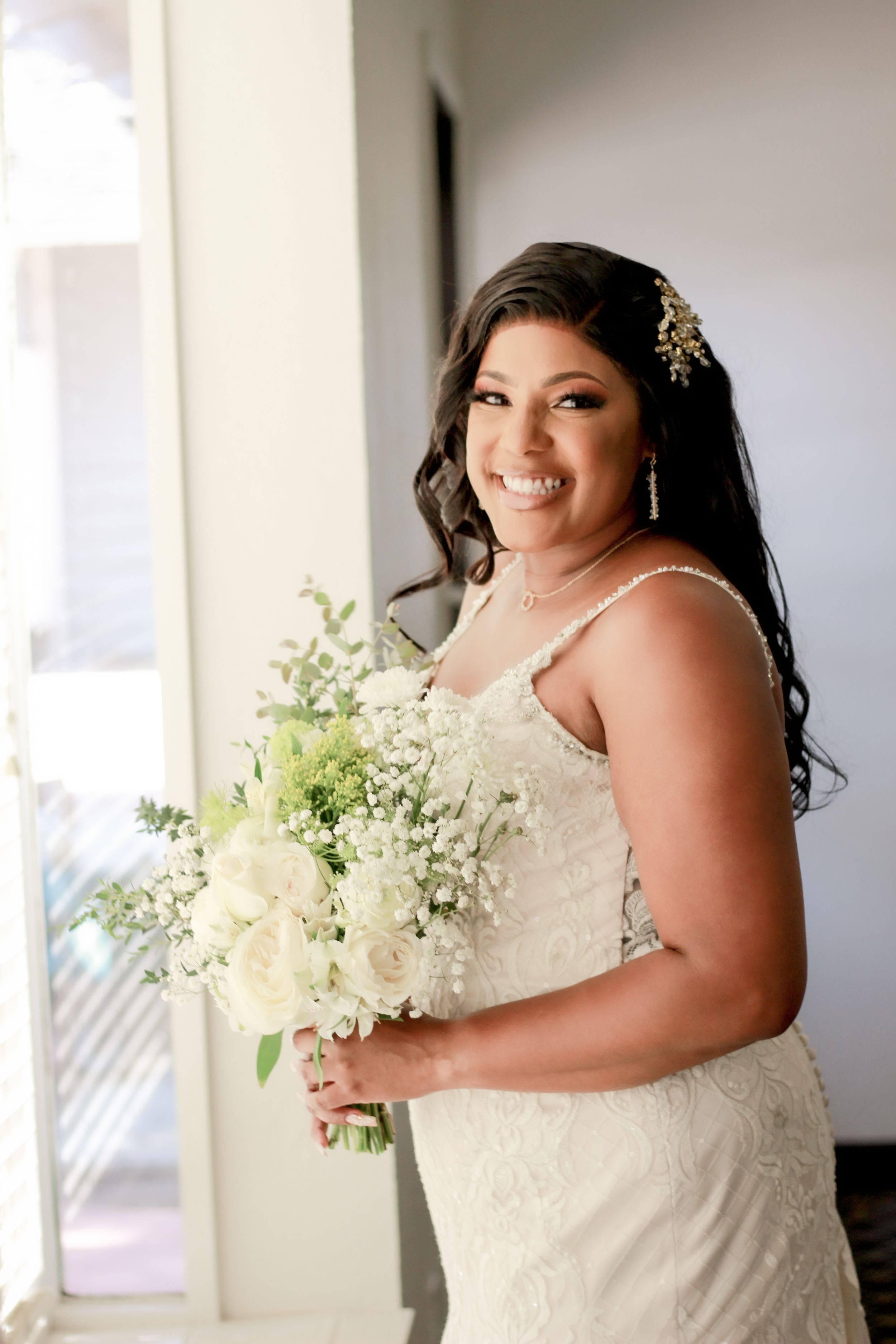 Bride holding white floral bouquet while standing by window in lace wedding dress.