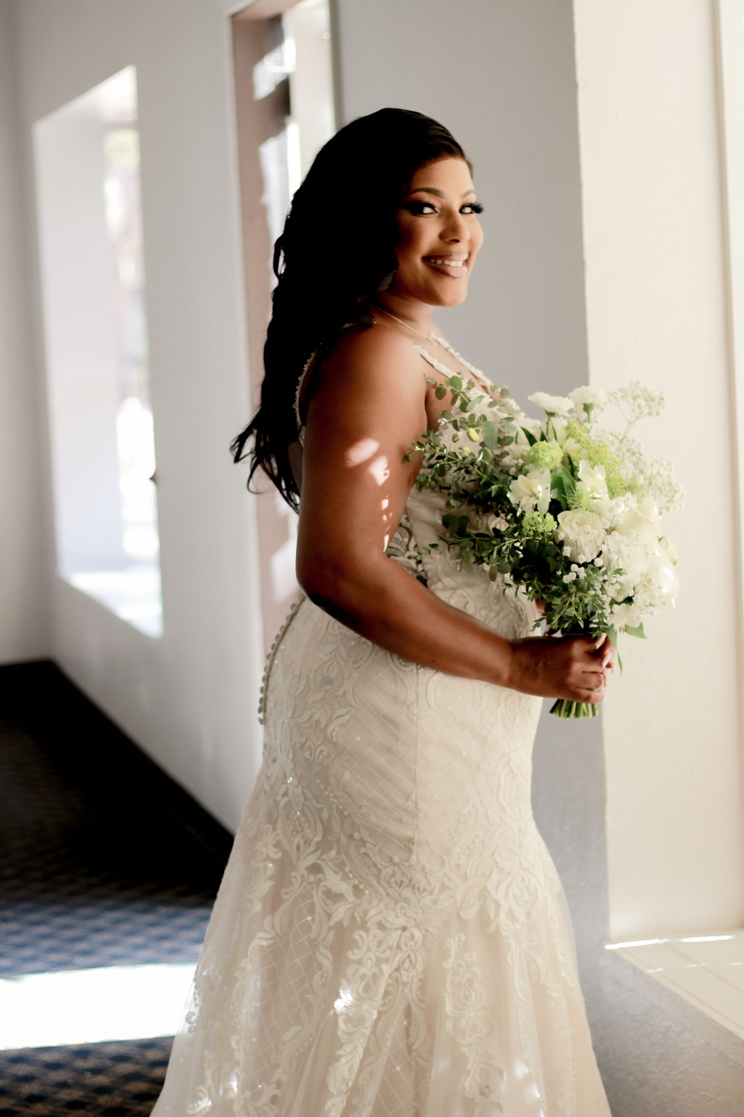 Bride in fitted lace wedding gown holding white and green bouquet in sunlit room.