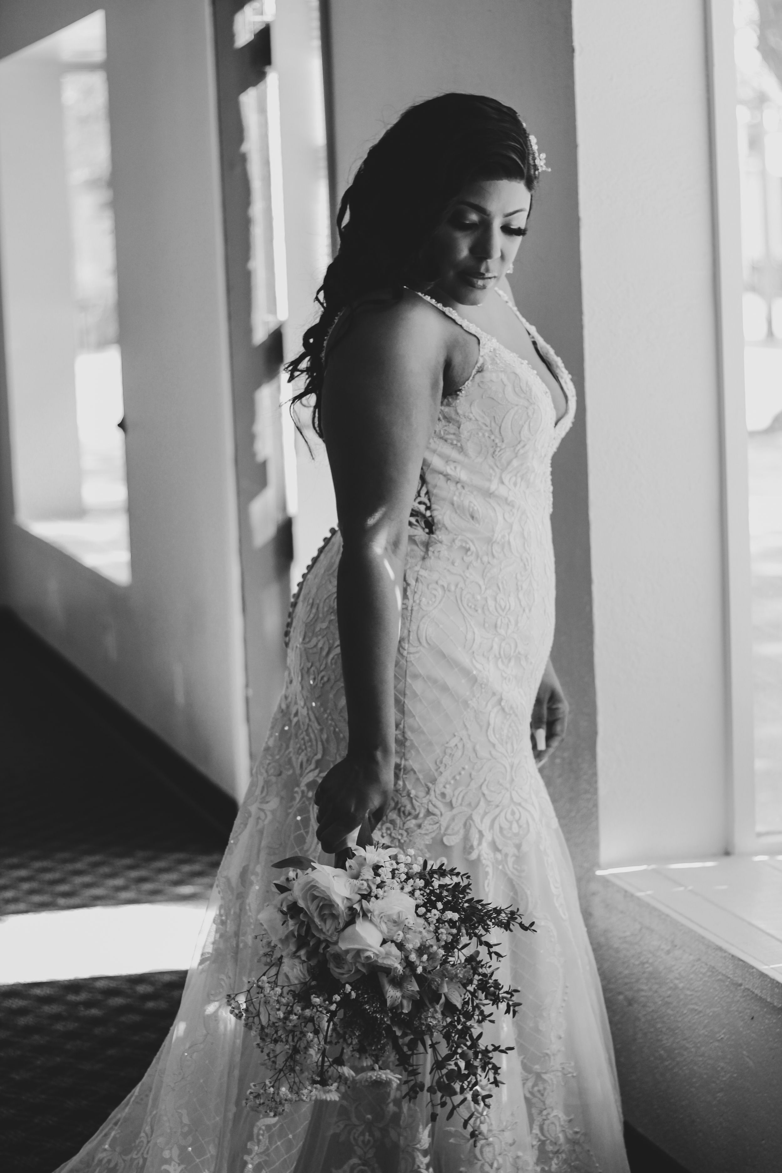 A bride in a lace wedding dress poses near sunlit windows in black and white.