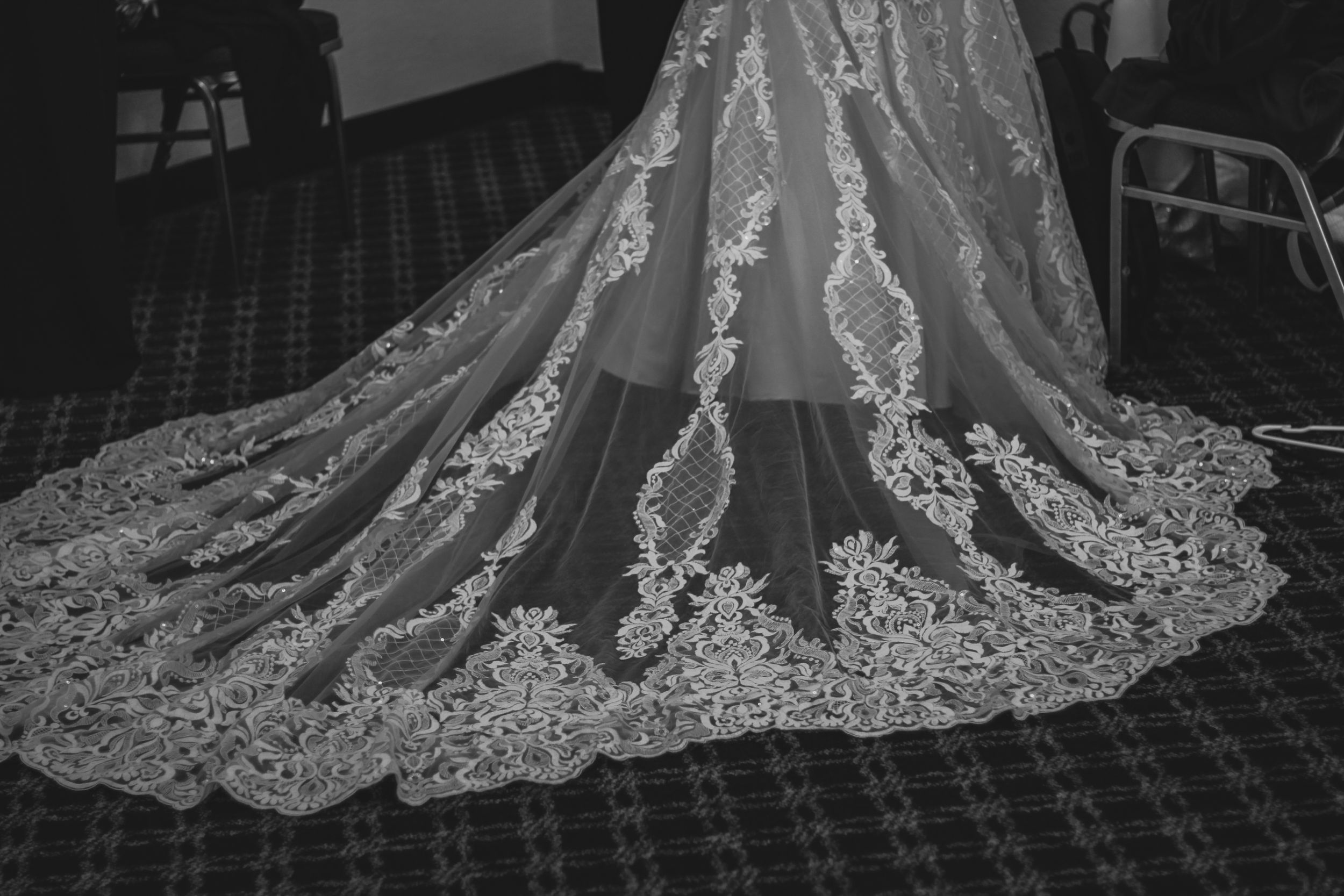Elegant lace train of a wedding dress spread out on patterned carpet in dramatic lighting.