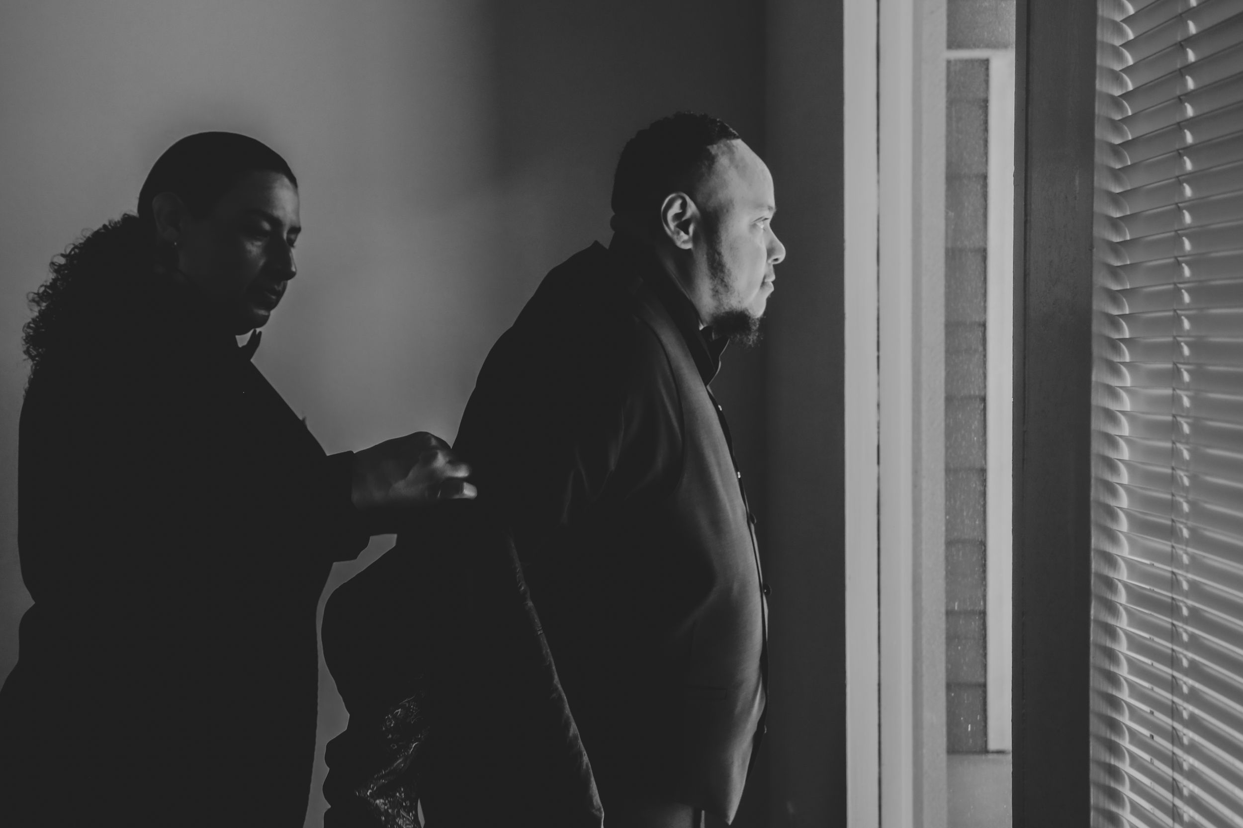 Two people standing by a window with venetian blinds in black and white.