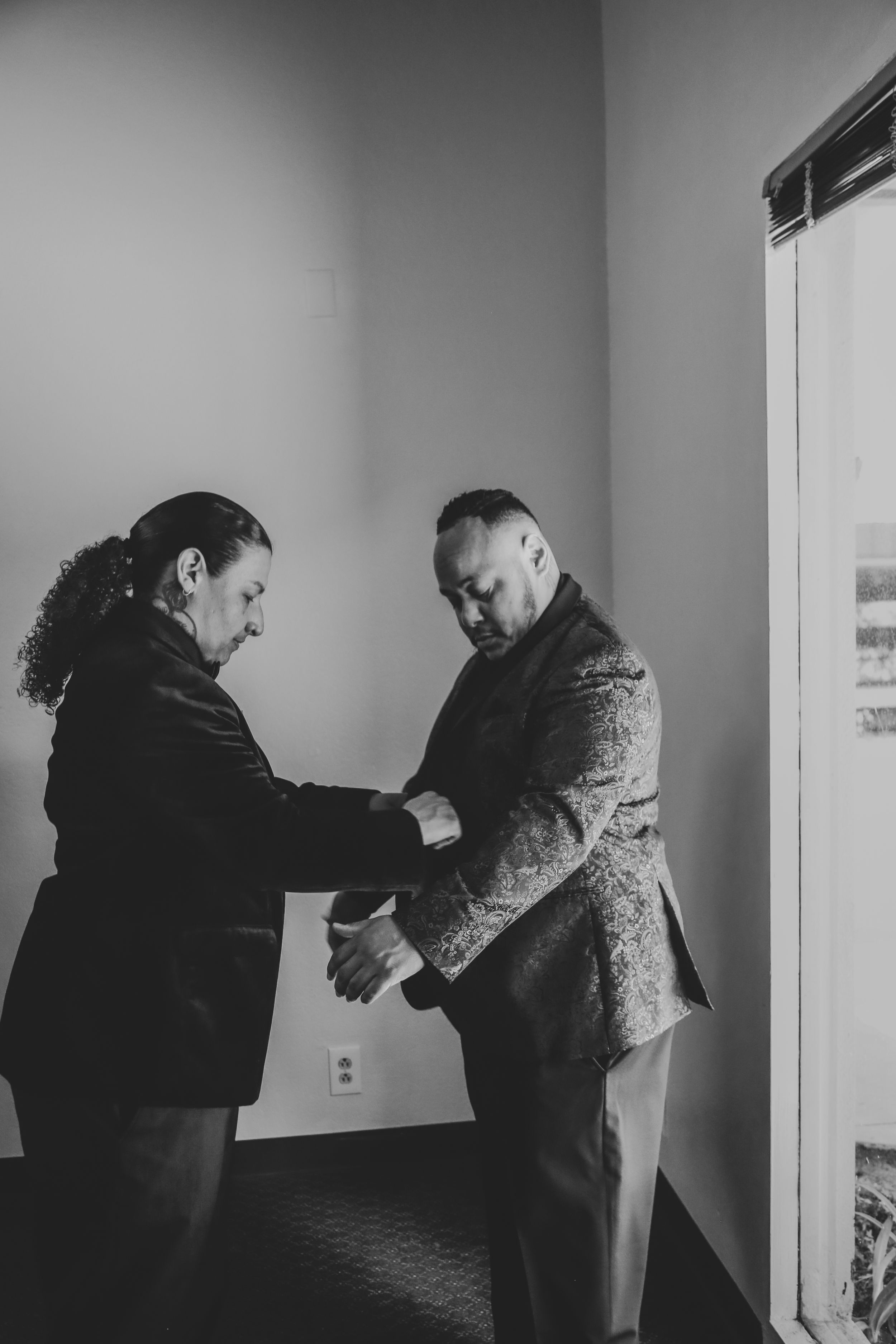 Two people helping with formal attire adjustments in a room.