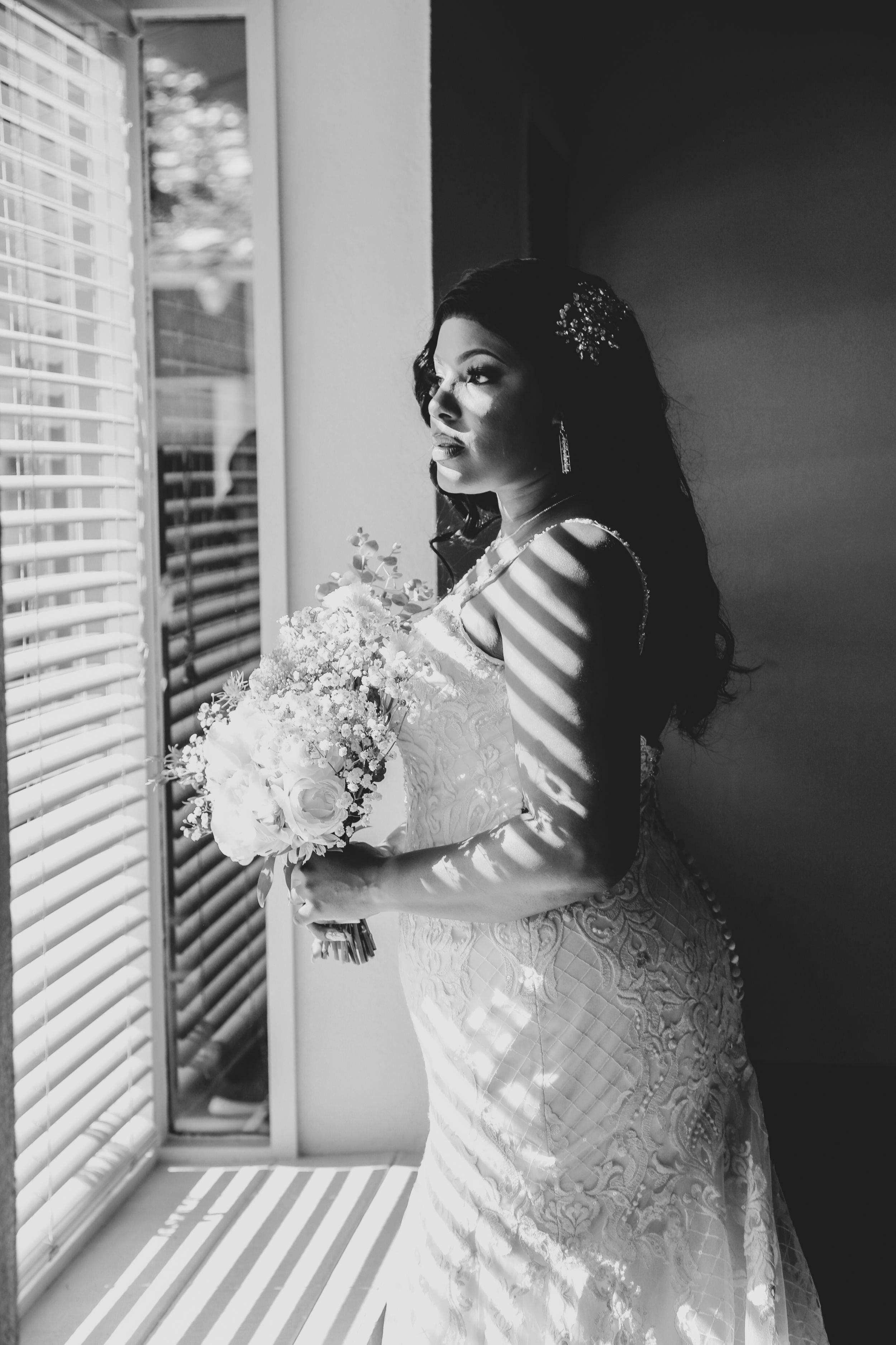 Bride in lace dress holding white bouquet by window in dramatic black and white.