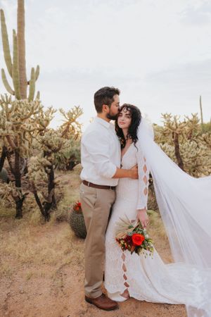 Bride in flowing white dress and groom in desert wedding attire embrace among desert plants.