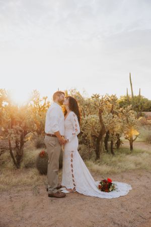 Newlyweds embrace in a desert garden during golden hour with a white lace dress and red bouquet.