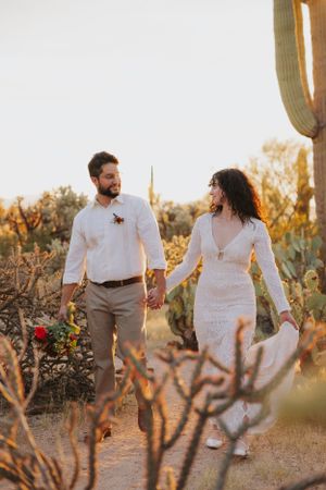 A couple strolls hand in hand through a desert landscape with tall saguaro cacti at sunset.