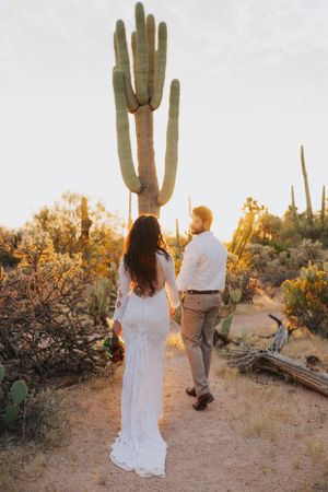 A couple in white clothing walks along a desert trail surrounded by majestic saguaro cacti.