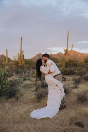 Couple embraces in a desert setting with towering saguaro cacti and mountains in the background.