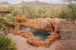 Desert landscape with red rock formations, mountains, and a natural pool surrounded by desert vegetation.