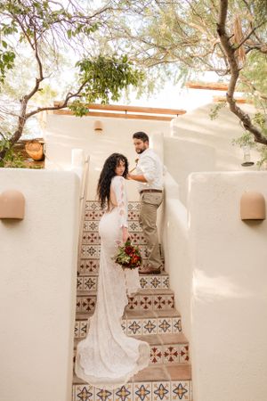 Wedding couple poses on decorative tiled steps against white adobe wall with hanging plants.