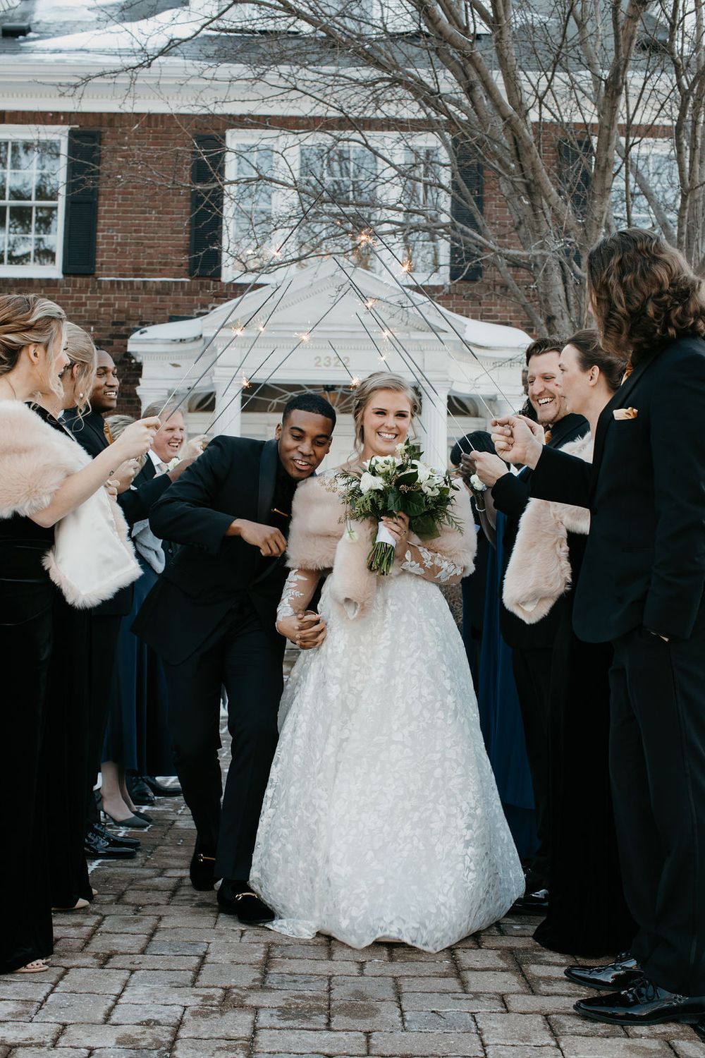 Smiling bride and groom walk through sparkler send-off with wedding party outside brick venue.