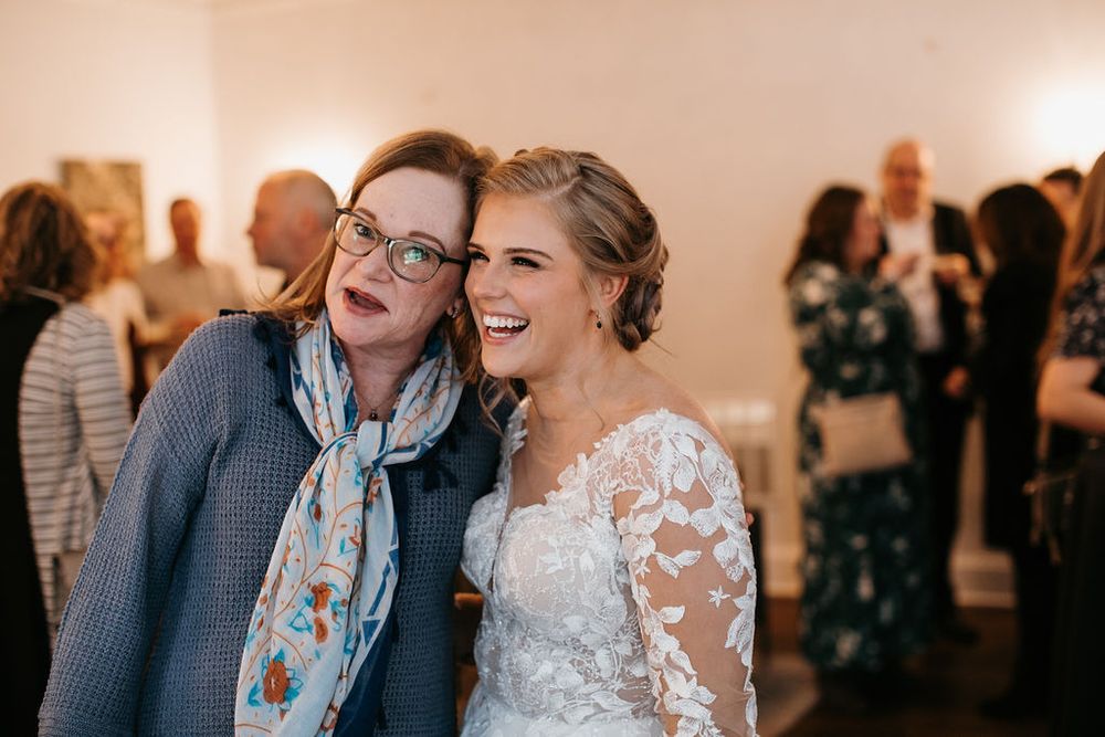 Bride laughs joyfully with a female guest wearing glasses at wedding reception.