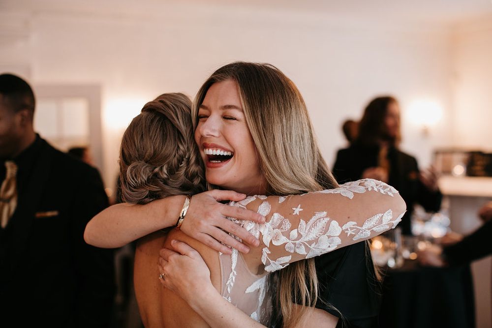 Bride in lace gown shares warm embrace with smiling female guest at reception.