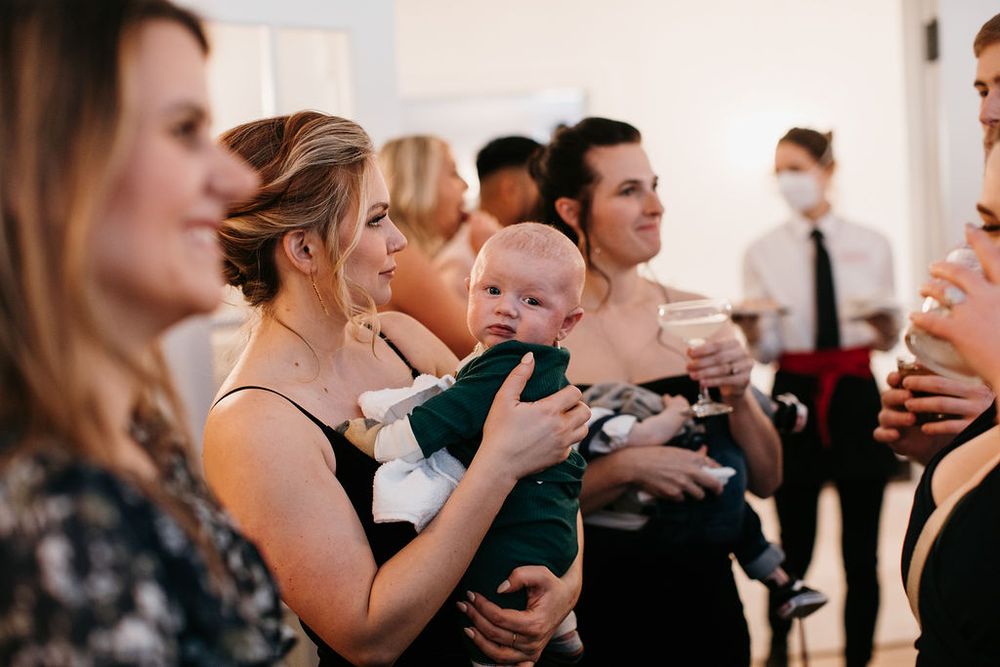 Wedding guest holds dressed-up baby in green outfit during indoor reception gathering.