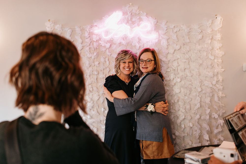 Two women share a warm hug in front of a floral backdrop with a pink neon light at a wedding reception.