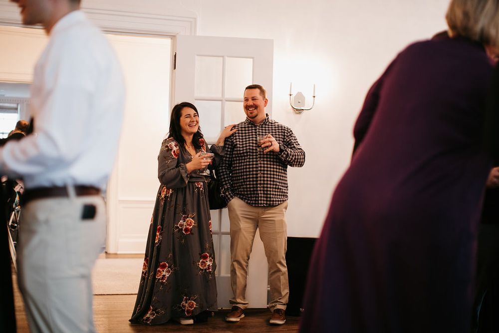 Smiling couple in floral dress and casual attire enjoy drinks at wedding reception.