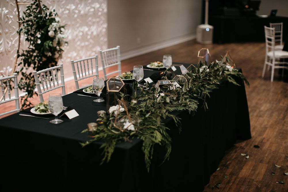 Elegant wedding reception head table with black linen, lush greenery garland, and gold candle holders.