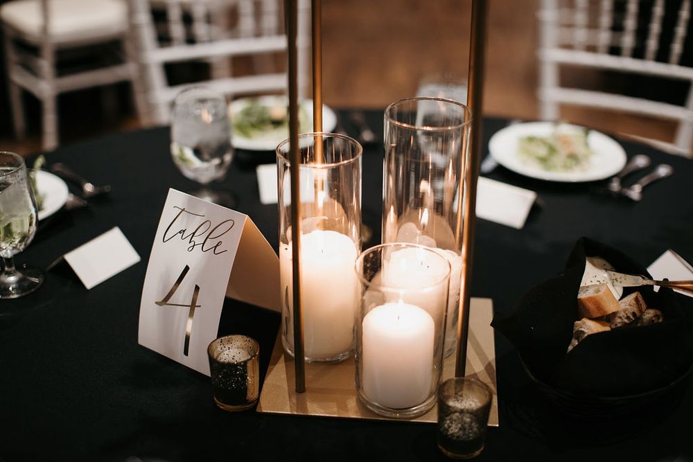 Gold geometric candle holder centerpiece with white pillar candles on a black tablecloth at Table 4.