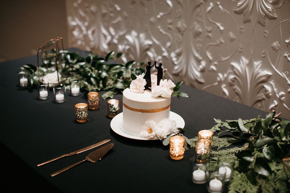 Simple white wedding cake with bride and groom topper surrounded by candles and greenery on black table.