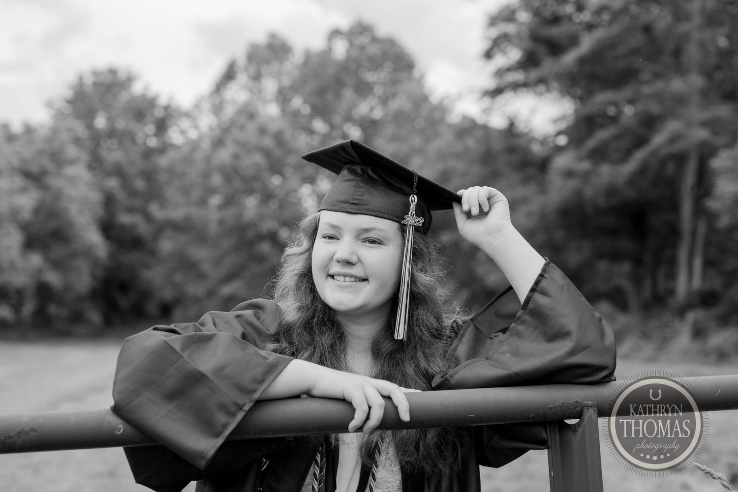Black and white portrait of graduate in cap and gown leaning on railing outdoors.