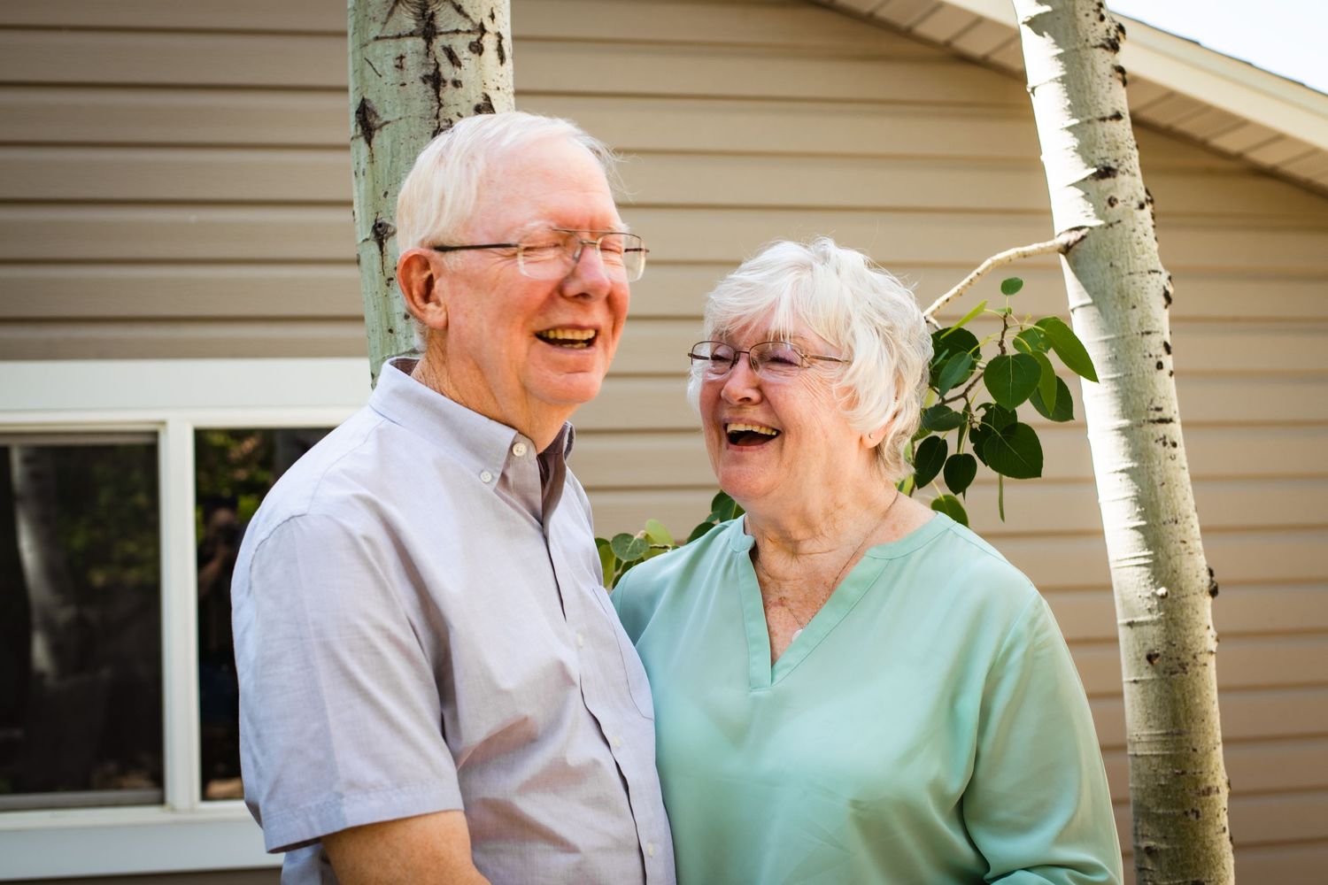 Elderly couple sharing genuine laughter outside their home with birch trees.