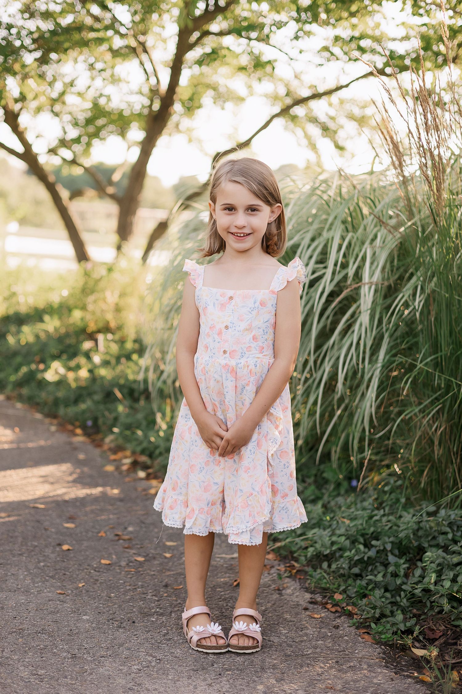 Child in floral dress stands on garden path near ornamental grasses.