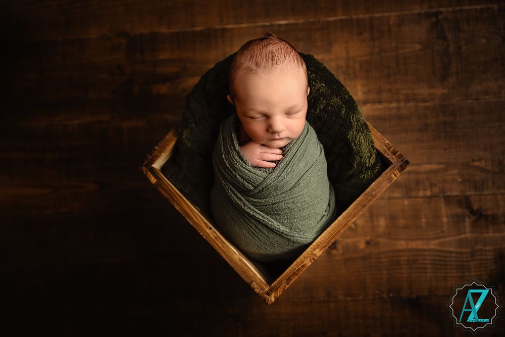 Top-down view of peaceful newborn in green wrap nestled in rustic wooden box against dark backdrop.