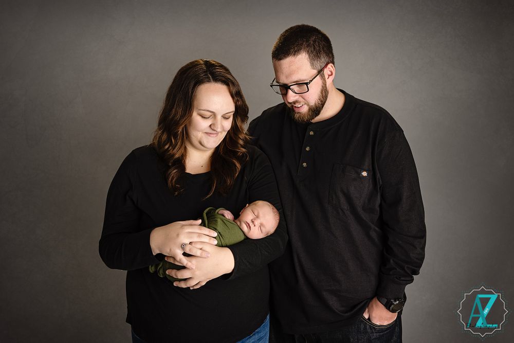 Studio portrait of new parents in black attire gazing lovingly at their sleeping newborn.