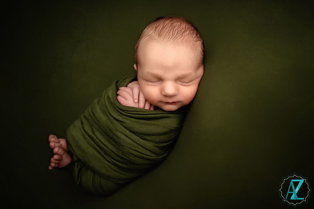 Newborn wrapped in olive green fabric sleeping peacefully against dark green background.