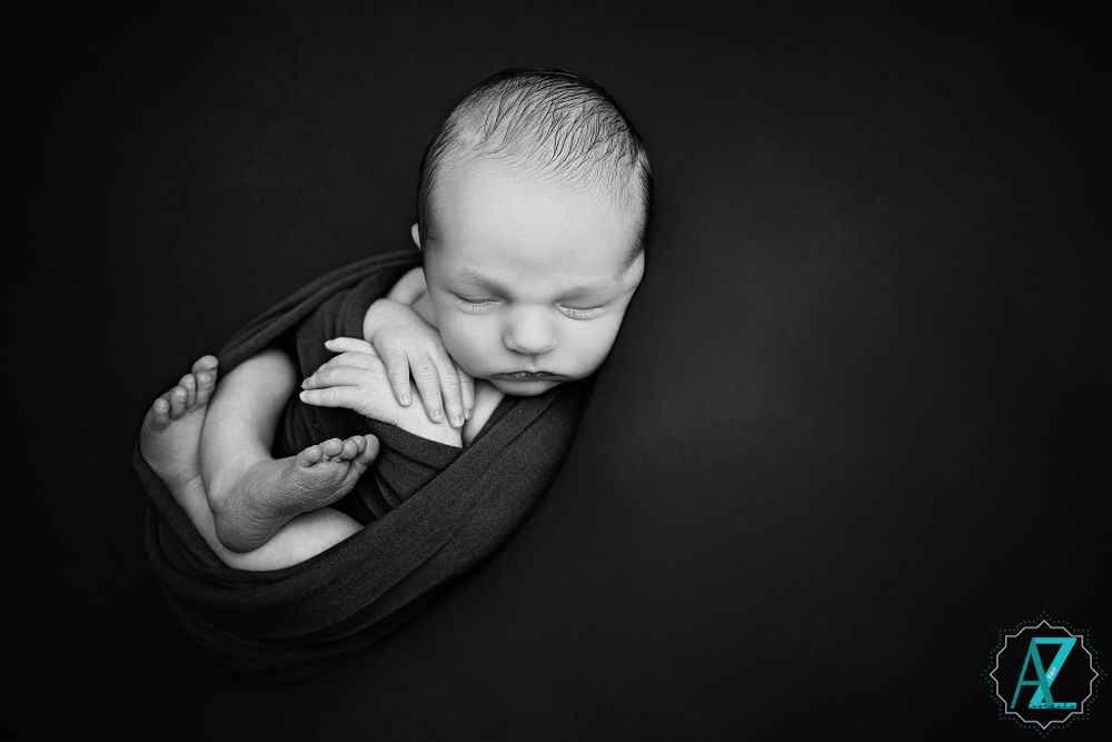 High contrast black and white portrait of sleeping newborn curled in dark fabric.