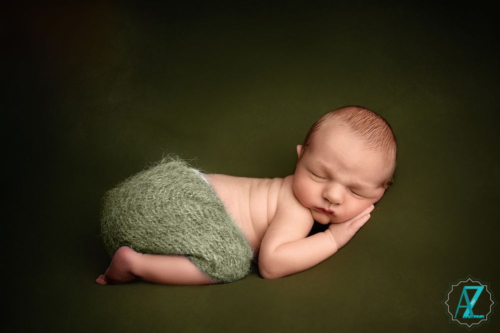 Artistic portrait of sleeping newborn wearing sage knit shorts against dark green backdrop.