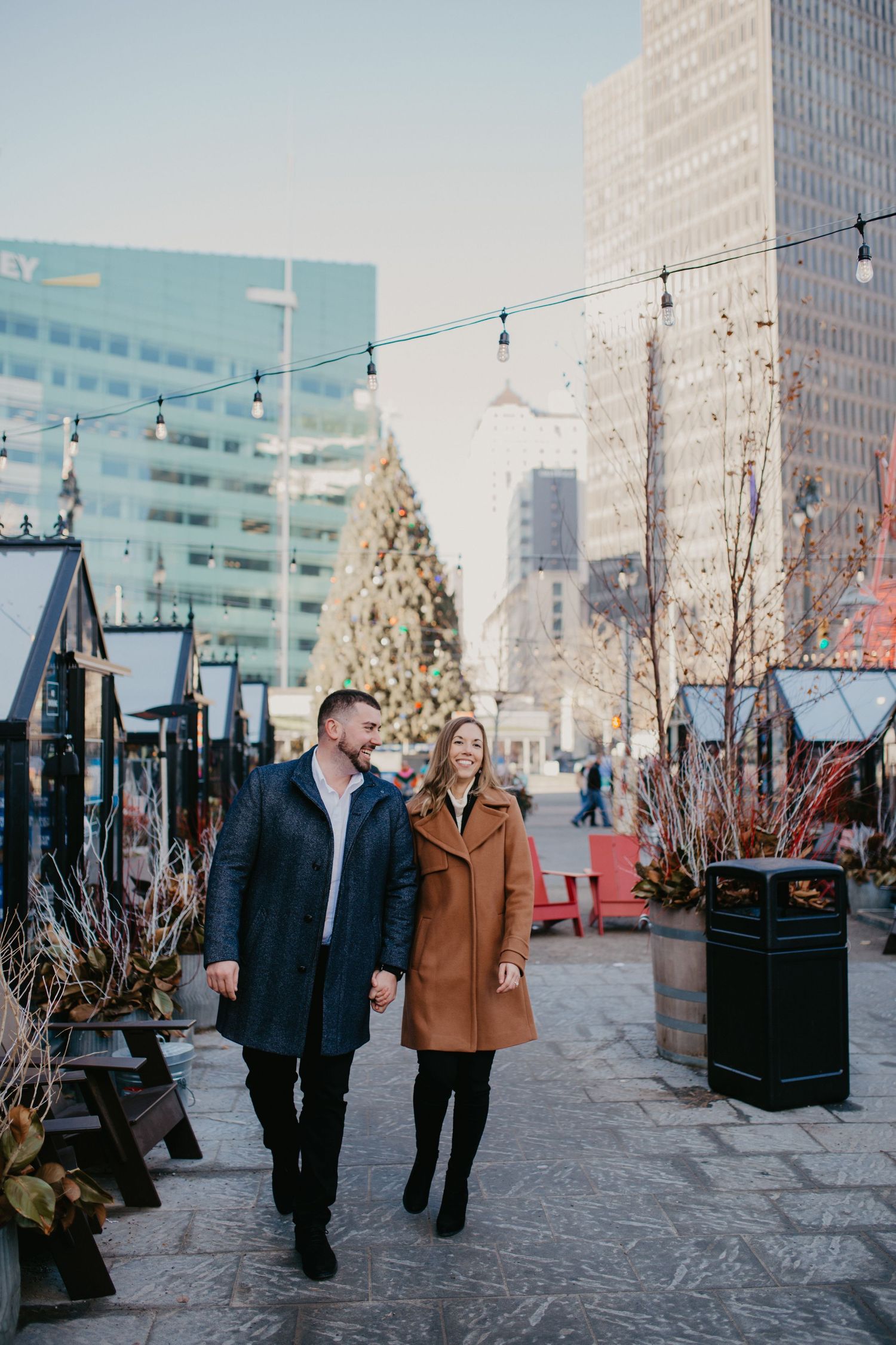 A couple strolls through a festive winter cityscape with string lights and holiday decorations.