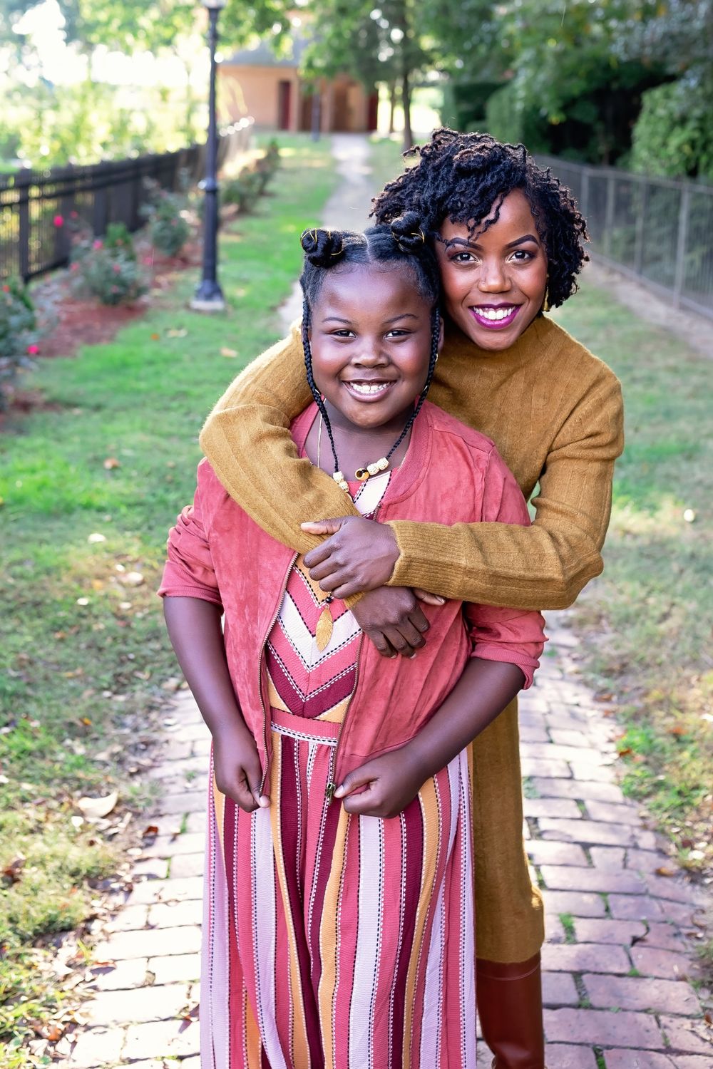 Woman hugging a young girl from behind on a brick pathway outdoors.