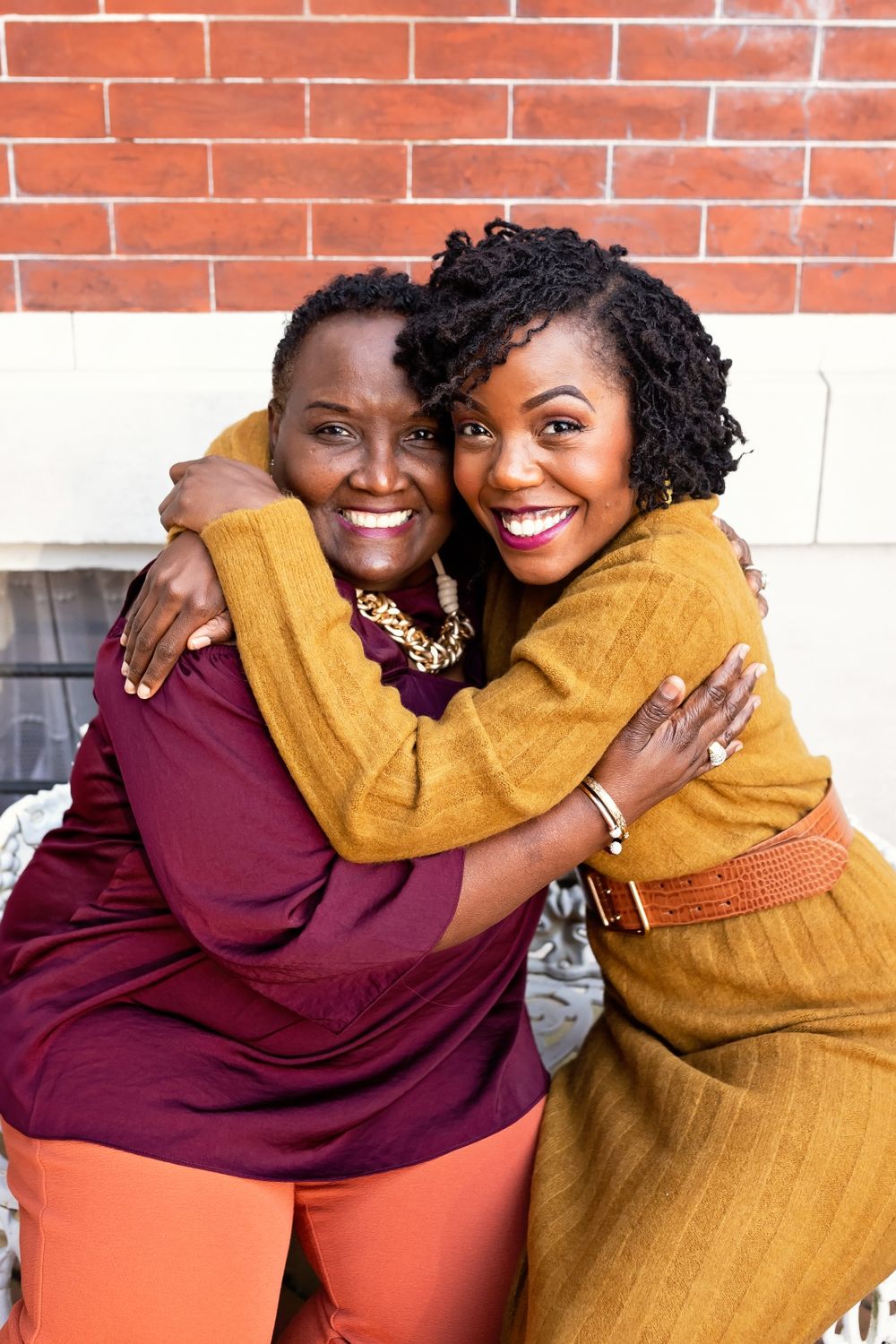 Two women sharing a warm hug and smiling outdoors near a brick wall.