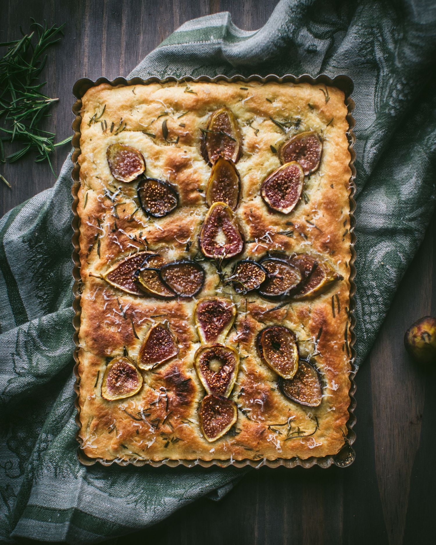 Golden focaccia bread topped with fresh figs and rosemary in a rectangular baking pan on a rustic wooden surface.