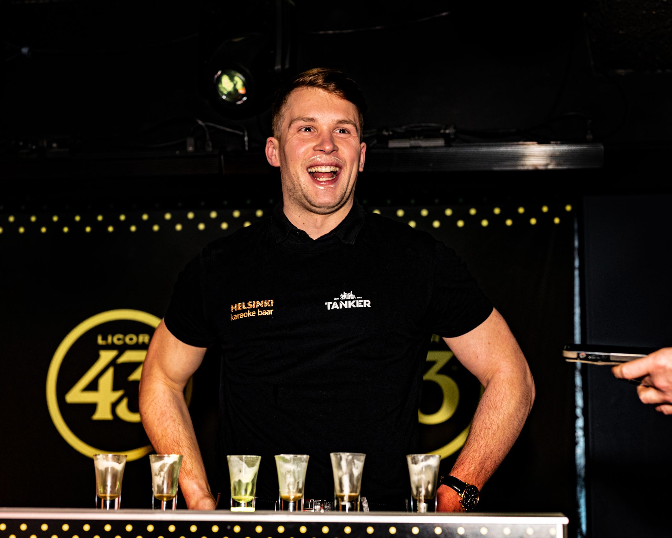 Energetic bartender performing flair moves behind bar counter with Licor 43 branding visible.