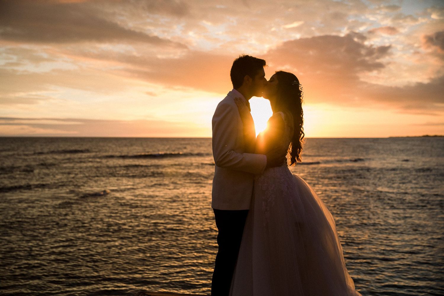 A silhouetted couple shares an intimate moment against a dramatic orange sunset over the ocean.