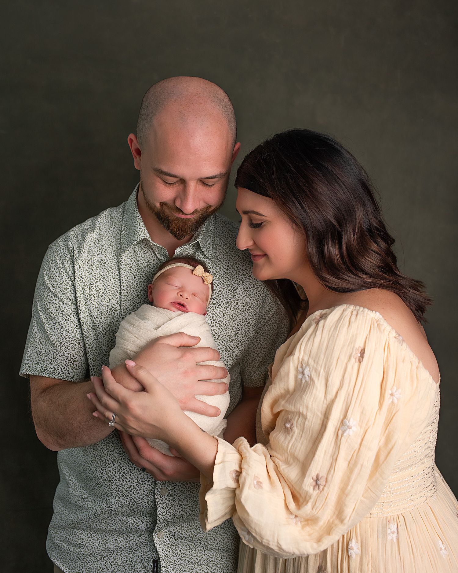 Proud bald father and brunette mother lovingly gaze at swaddled newborn baby in studio portrait.