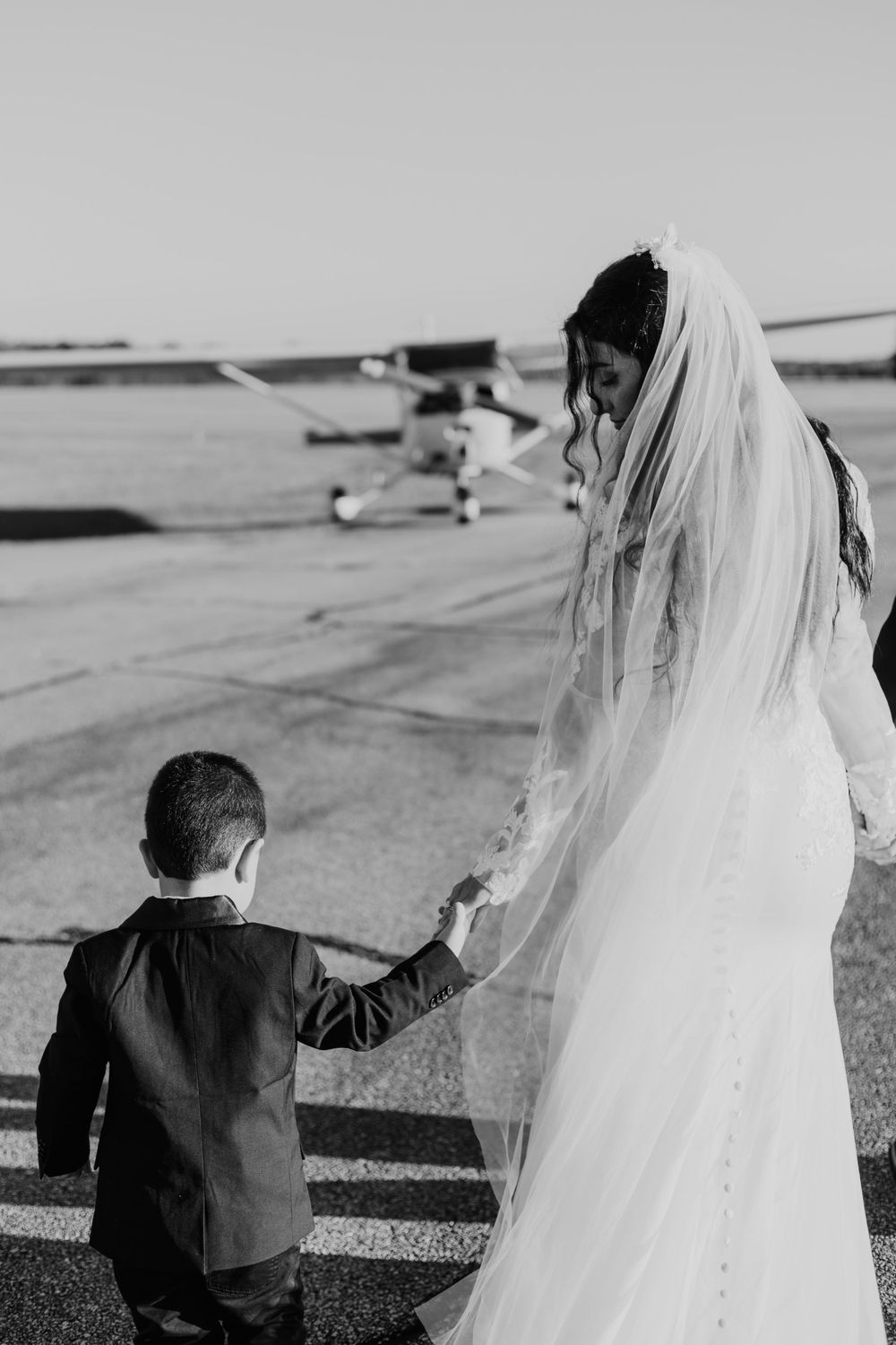Black and white photo of a bride and child walking near airplanes at an airfield.