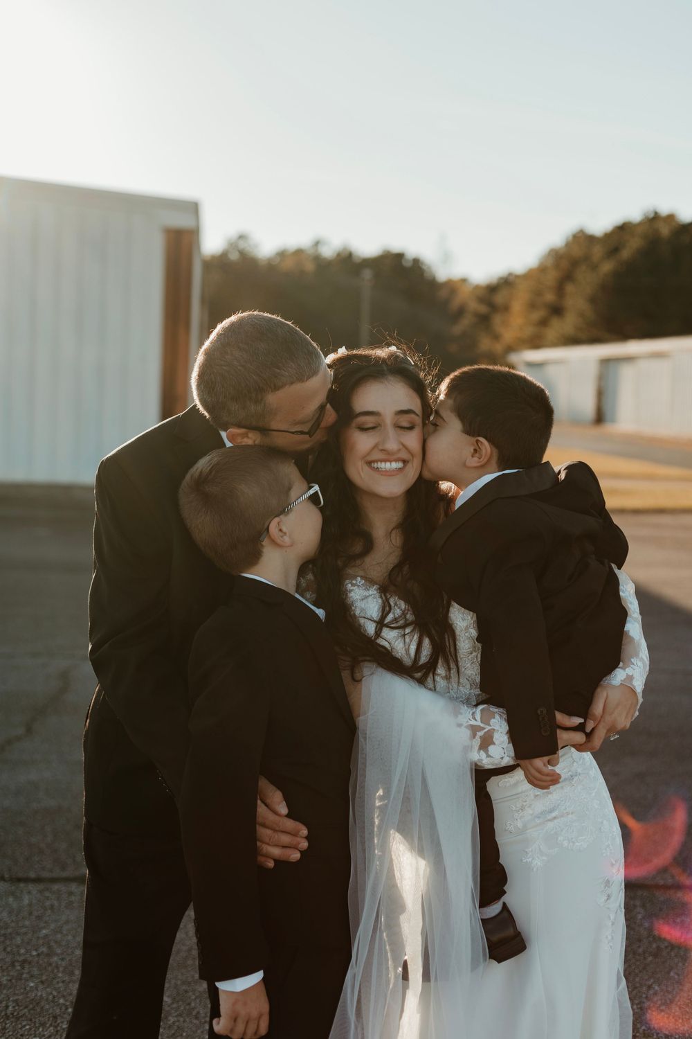 Sweet moment of family members kissing bride on cheeks during sunset outdoor photo session.