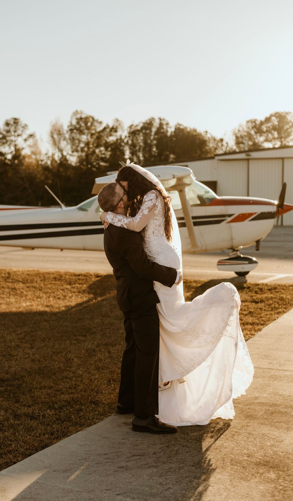 A romantic wedding moment at sunset with a small airplane in the background at an airfield.
