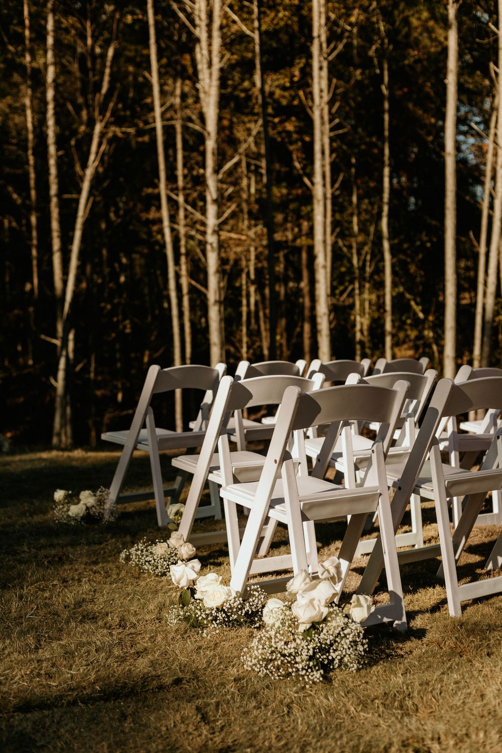 White folding chairs lined up for an outdoor forest wedding with floral aisle decorations.
