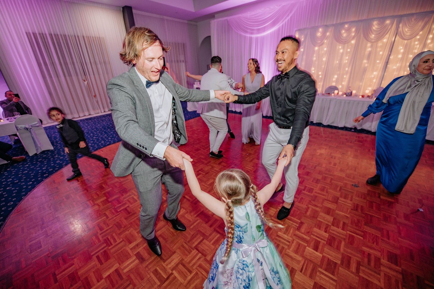 People dancing together at a wedding reception on a red wooden dance floor with purple lighting.