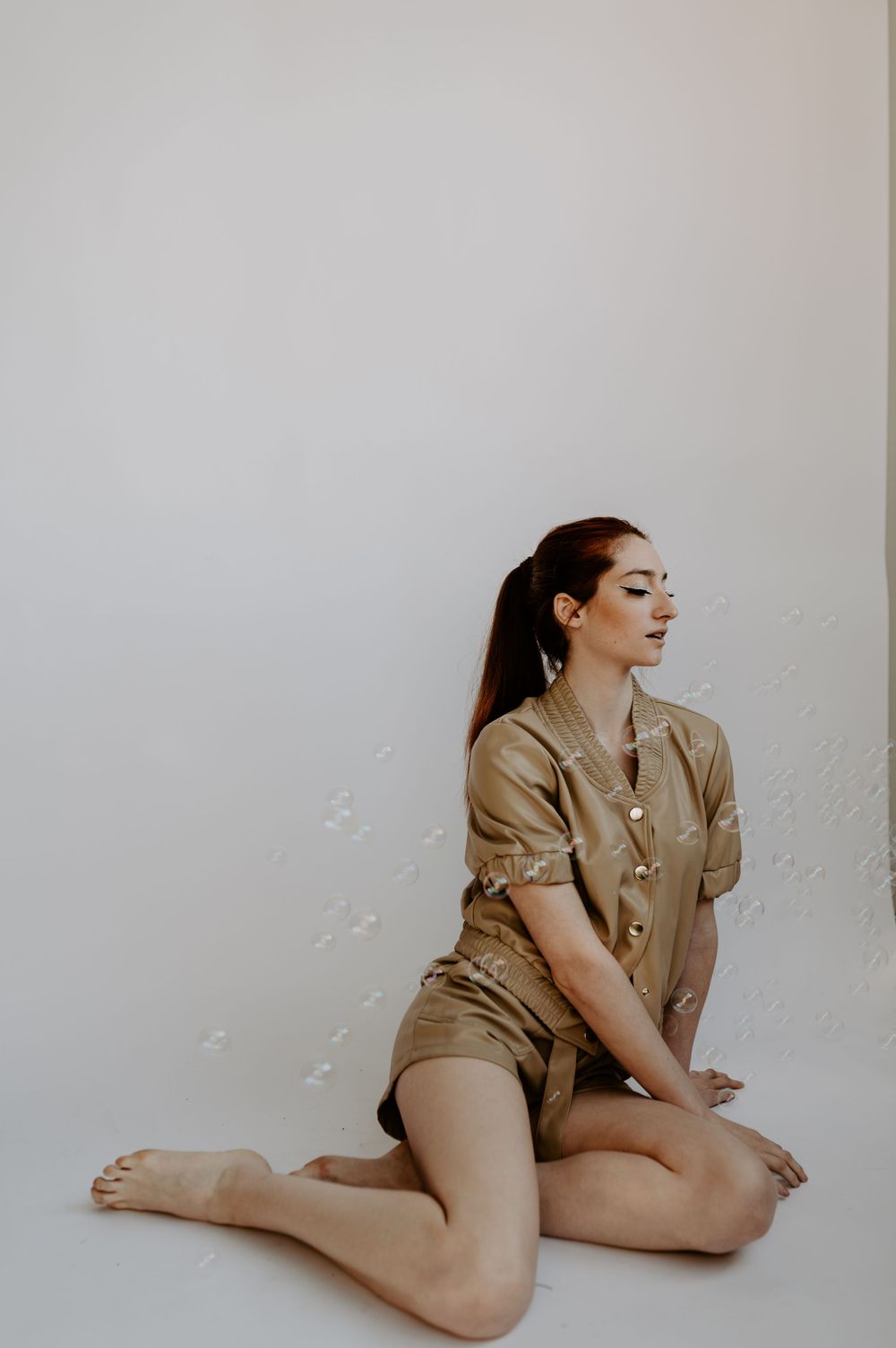 Minimalist studio portrait of person in beige outfit against white wall.